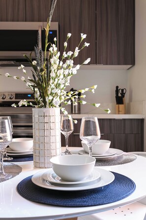 A modern dining table set with white bowls and wine glasses, a decorative vase with white flowers, and a kitchen in the background. - The Penn Hotel - Smart TV & King Bed Near Gaslamp! (San Diego)