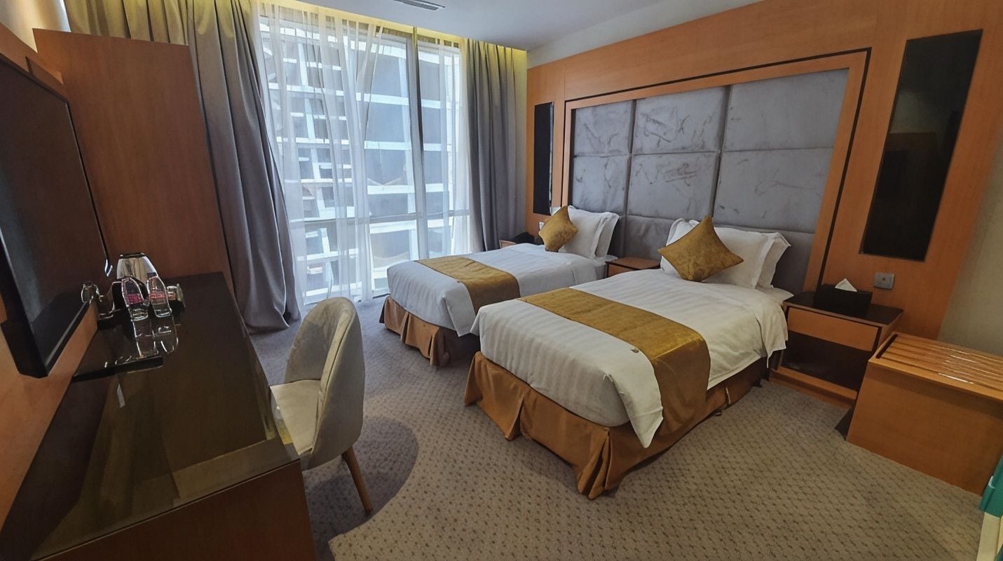 Habitación de hotel con dos camas, un escritorio, una silla y un amplio ventanal con cortinas.