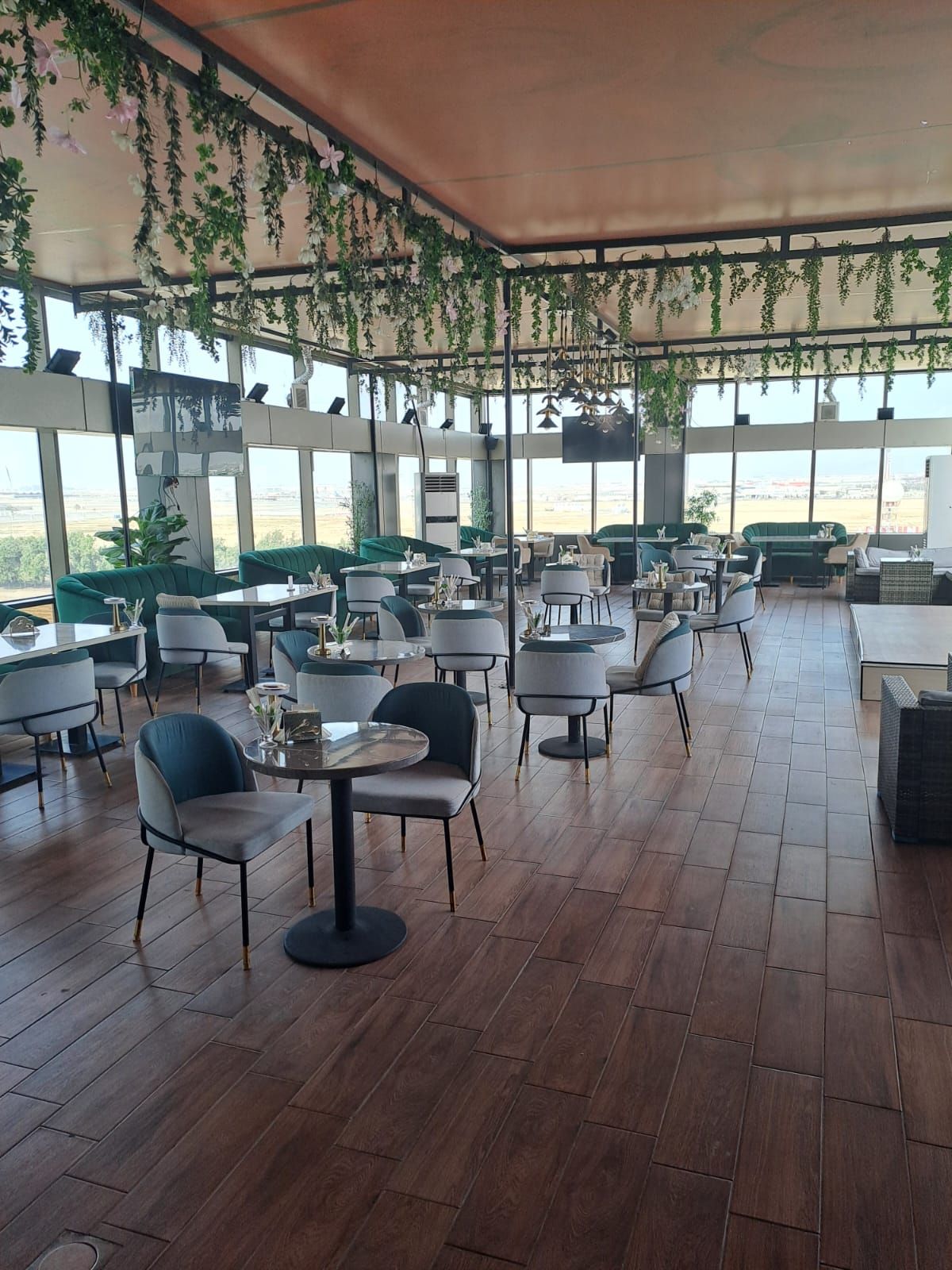 Un comedor interior con varias mesas y sillas, ventanas amplias y plantas colgantes.