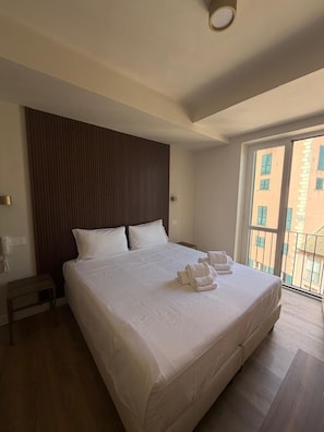 Habitación de hotel con una cama grande, cabecera de madera, mesita de noche y vistas a edificios a través de una ventana.