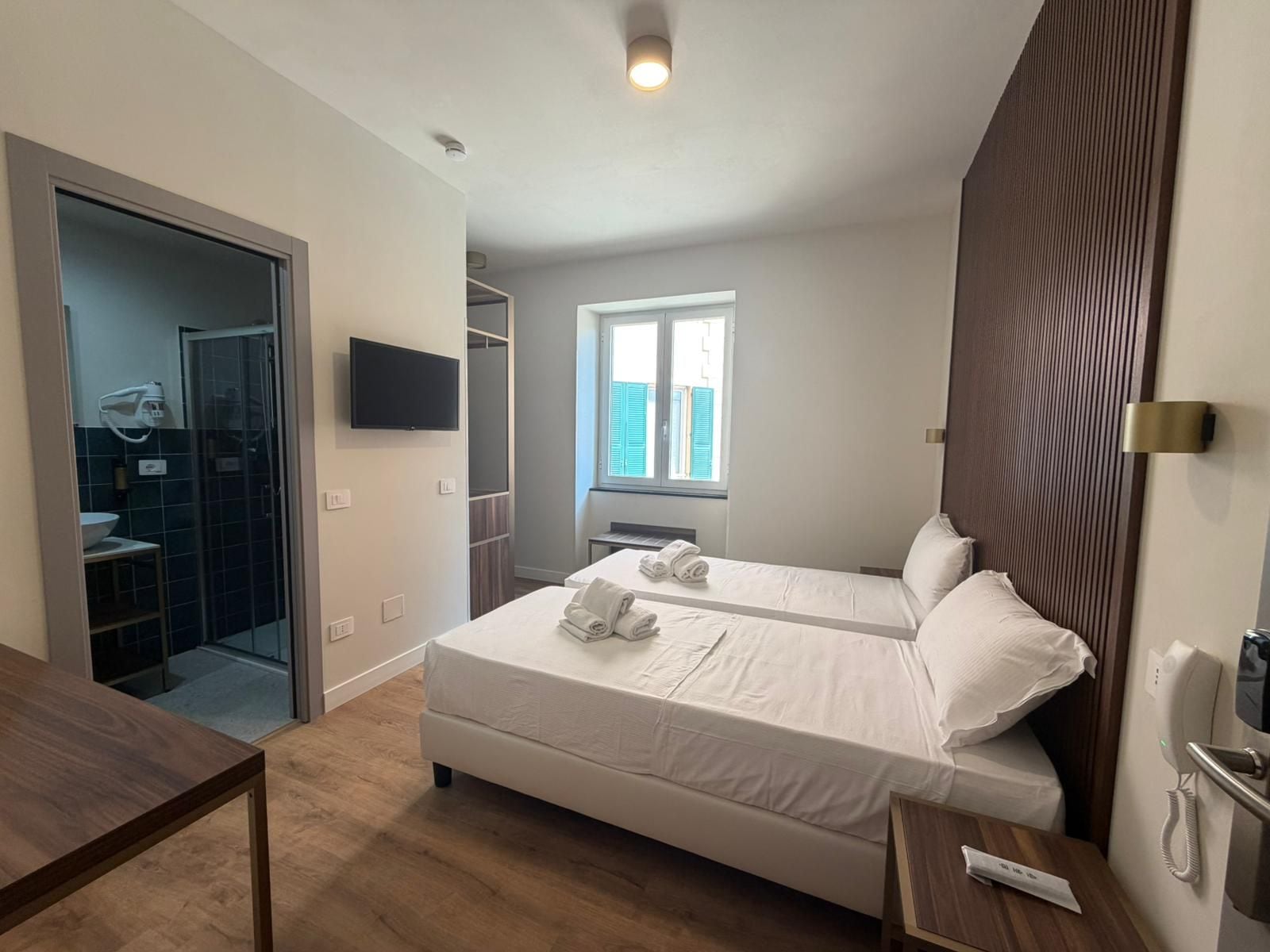 A hotel room with two beds, a TV, a bedside table, a window, and a bathroom with a shower and sink.