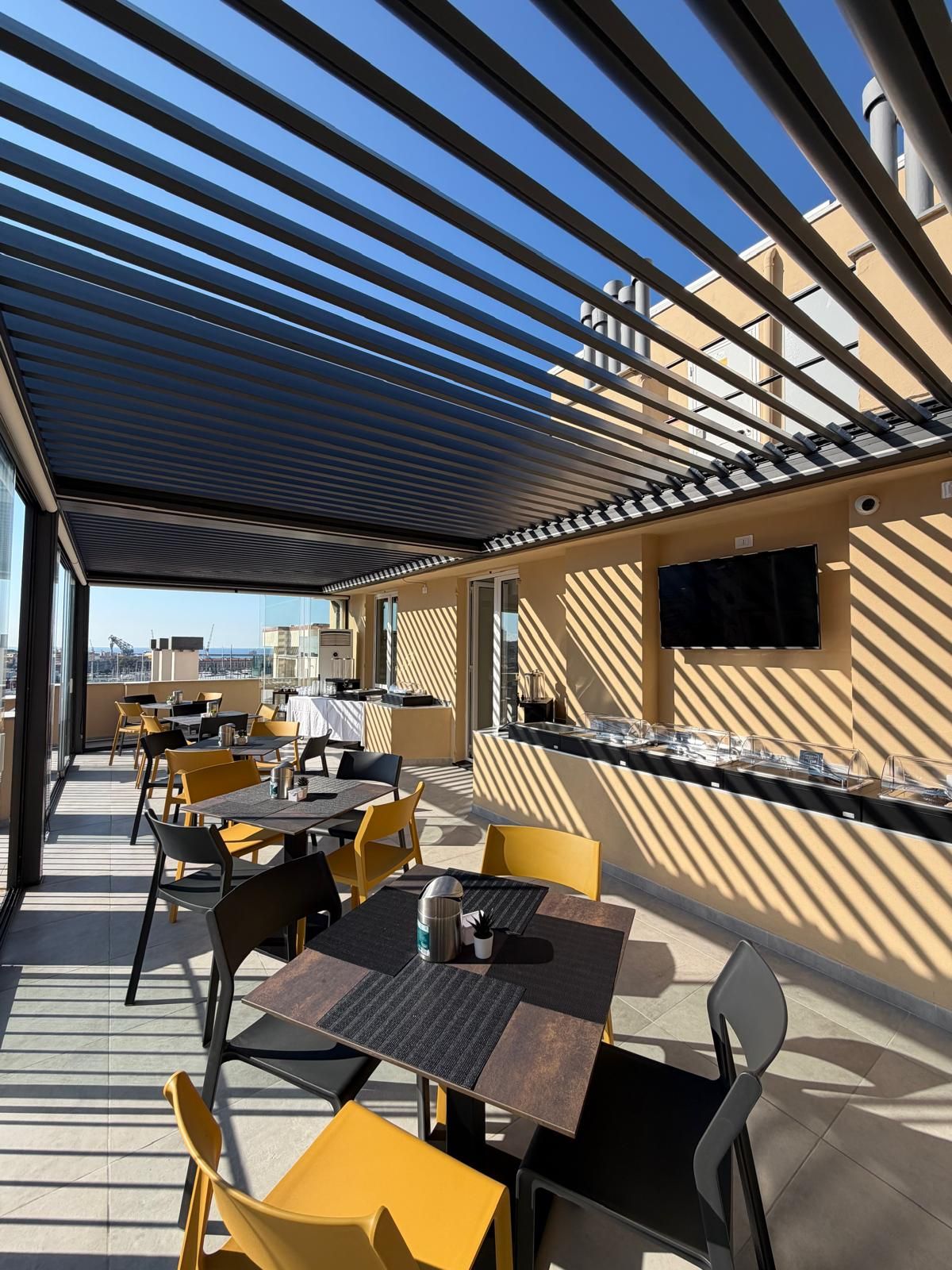 Un patio al aire libre con mesas y sillas, un televisor de pantalla plana montado en la pared y vistas de la ciudad al fondo.