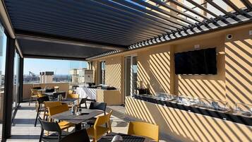 Un patio al aire libre con mesas y sillas, un televisor de pantalla plana montado en la pared y vistas de la ciudad al fondo.