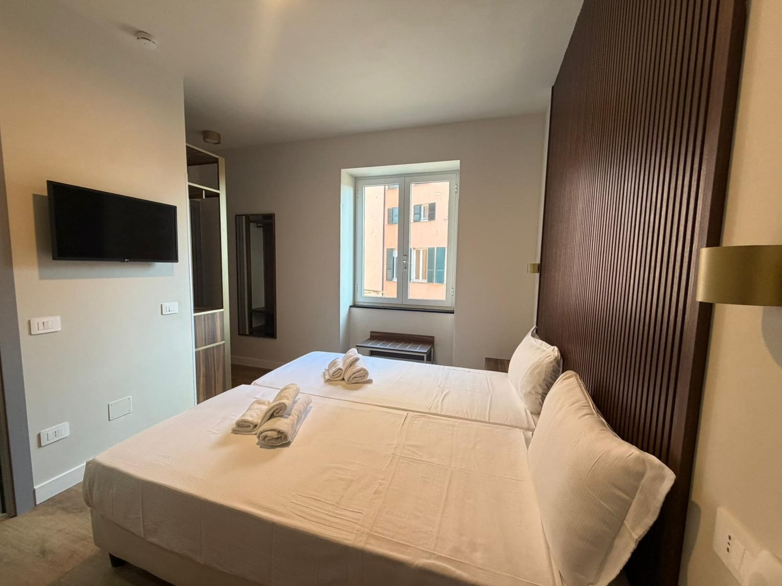 Een hotelkamer met een bed, een aan de muur bevestigde televisie, een raam met uitzicht op gebouwen en een deur naar een gang.