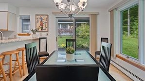 A dining area with a glass-top table, black chairs, a chandelier, and a view of a garden through large windows. - The Woods Killington - 2B with Spa, Pool & Jacuzzi (Killington)