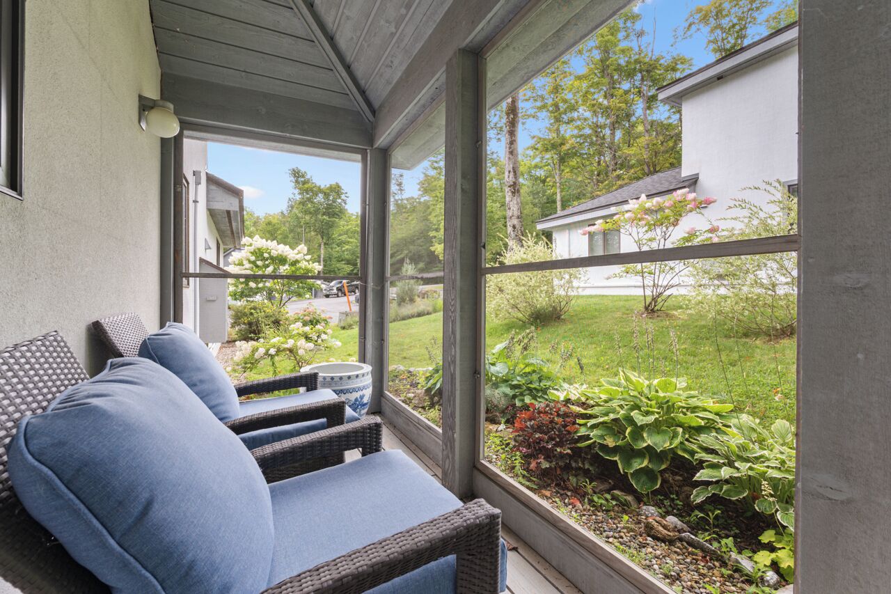 Área de estar coberta ao ar livre com mobiliário de vime e vista para um jardim e casas vizinhas.