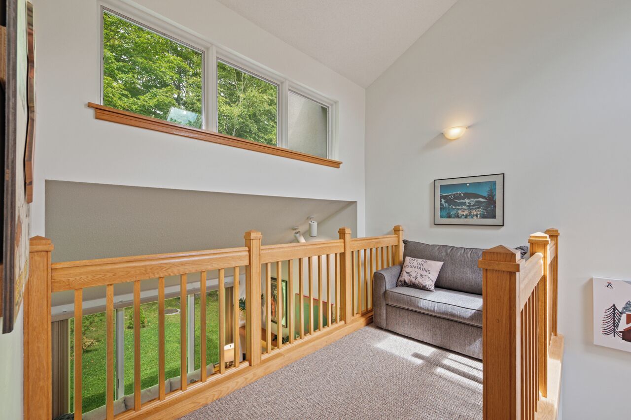 A room with a wooden railing, a grey sofa, a framed picture on the wall, and a window overlooking a green garden.