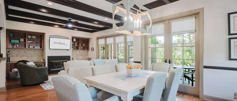 Amplia sala de estar con chimenea, comedor con mesa blanca y sillas grises, y cocina con gabinetes de madera.