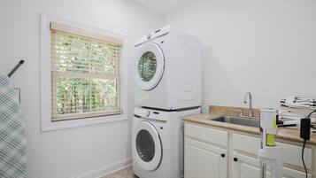 A laundry room with a stacked washer and dryer, a window with blinds, a sink, and a countertop with a towel rack.
