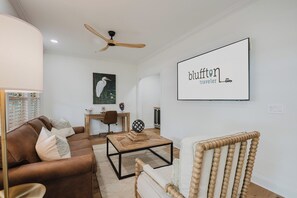 A modern living room with a brown leather sofa, a wooden coffee table, a wooden chair, a ceiling fan, and a framed artwork on the wall.