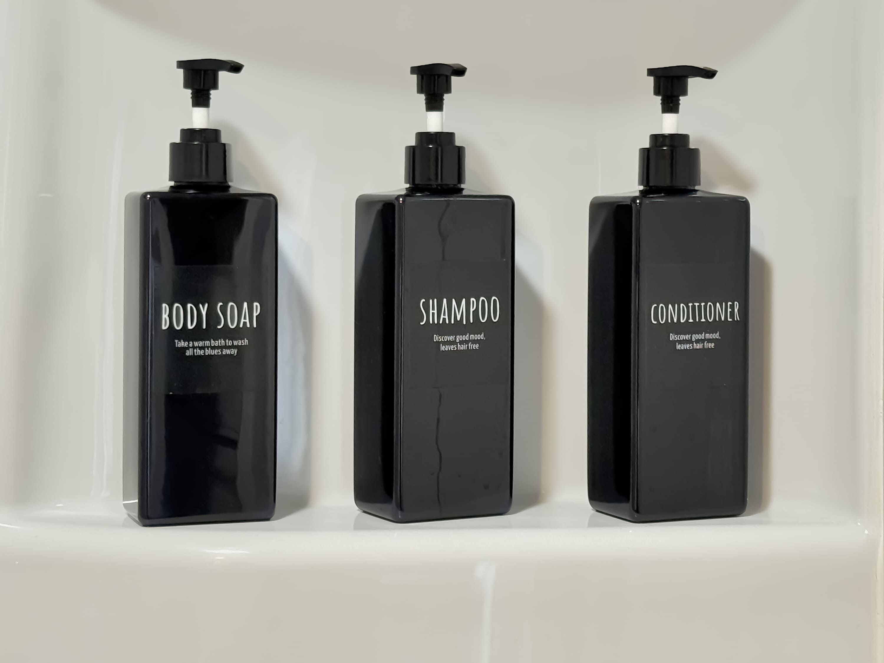 Three black bottles of body soap, shampoo, and conditioner on a white shelf.