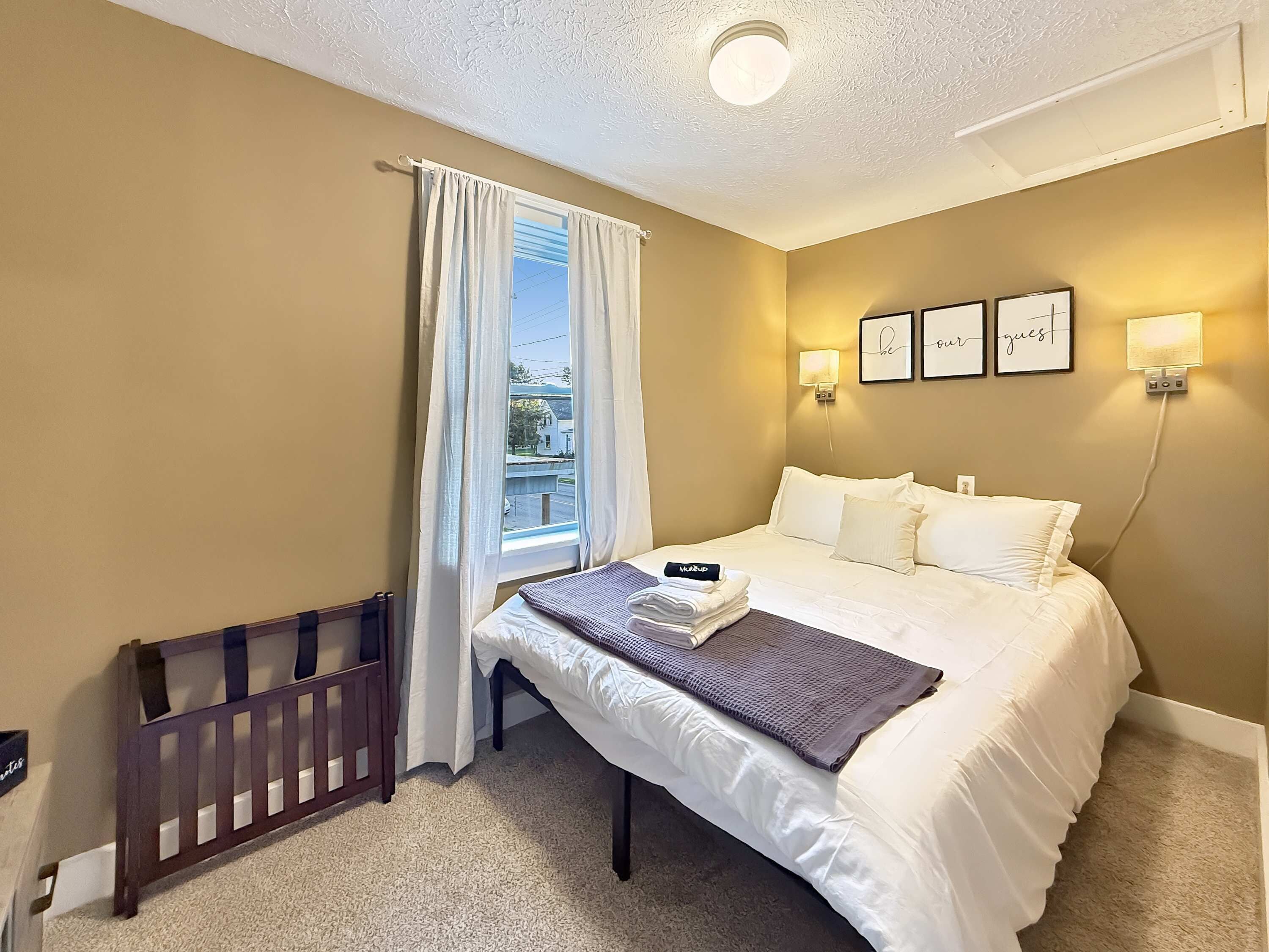 Habitación de hotel con cama, cuna, ventana con cortinas, luces fijadas en la pared y cuadros enmarcados.