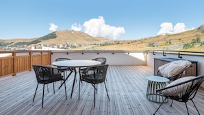 Um deck externo com mesa e cadeiras, com vista para uma paisagem montanhosa e edifícios ao longe.