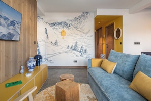 A modern living room with a large wall mural, a sofa, a wooden coffee table, and a yellow cabinet. - Solévivre – Terrace | Spa | View| Alpe d’Huez (Huez)