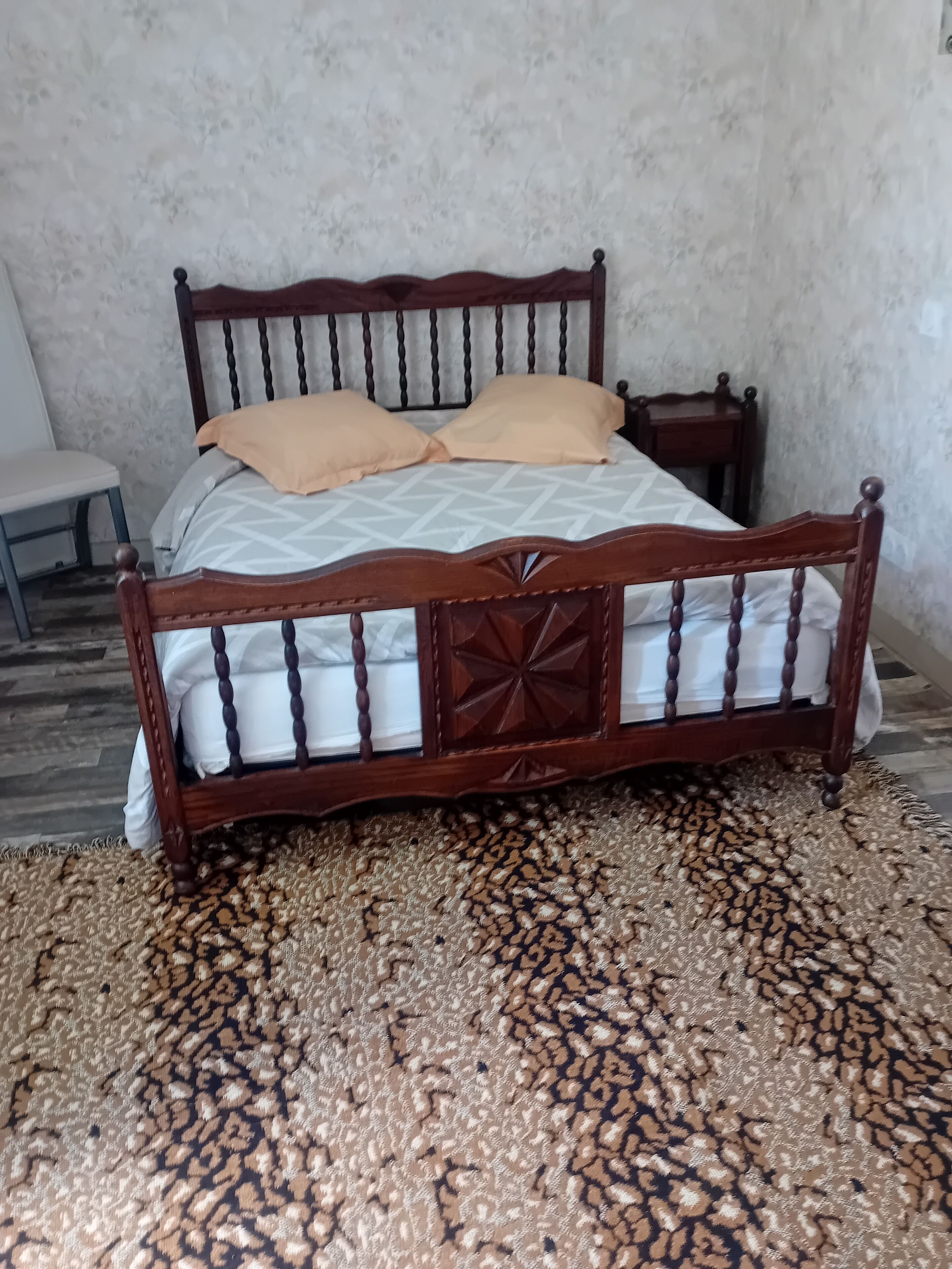 A bed with a wooden frame and a patterned bedspread, a small bedside table, and a chair.