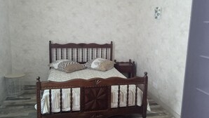 A single bed with a wooden frame and a patterned bedspread, positioned against a plain wall with a small round table beside it.