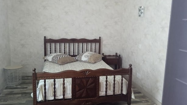 Un lit simple avec un cadre en bois et une couvre-lit à motifs, placé contre un mur uni, avec une petite table ronde à côté.