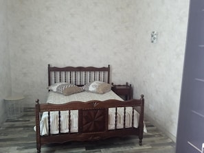 A single bed with a wooden frame and a patterned bedspread, positioned against a plain wall with a small round table beside it.