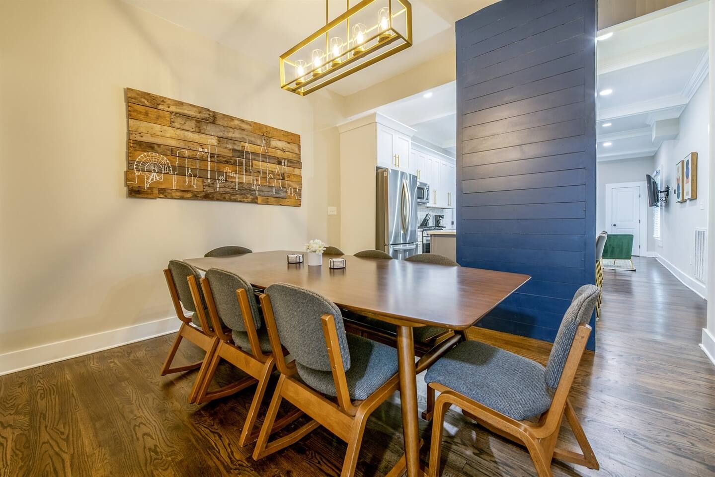 Un comedor con mesa y sillas de madera, una obra de arte con motivo urbano en la pared y una cocina moderna al fondo.