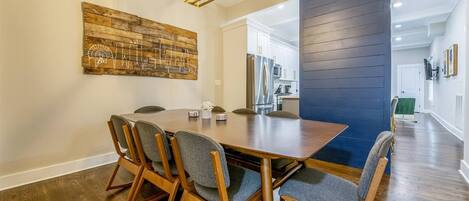 Un comedor con mesa y sillas de madera, una obra de arte con motivo urbano en la pared y una cocina moderna al fondo.