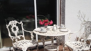 Un espacio de estar al aire libre con muebles blancos, sillas de metal ornamentadas y una mesa, ubicado frente a una ventana con vistas a una montaña.