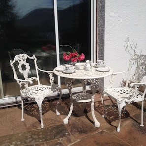 A white outdoor seating area with ornate metal chairs and a table, positioned in front of a window with a view of a mountain. - Apartment Sleeps 2 With Parking, Pets Welcome (Penrith)