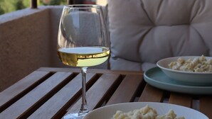 Ruokapöytä, jonka päällä on lasi valkoviiniä ja lautanen pastaa, pehmustettu tuoli ja taustalla näkyvä rakennusmaisema.
