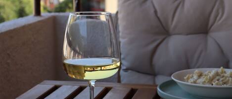 A dining table with a glass of white wine and a plate of pasta, a cushioned chair, and a view of buildings in the background.