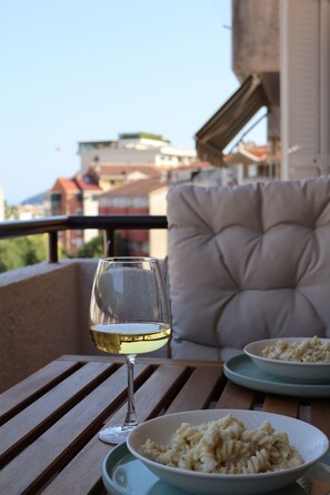 Mesa de jantar com um copo de vinho branco e um prato de massa, uma cadeira acolchoada e vista para edifícios ao fundo.