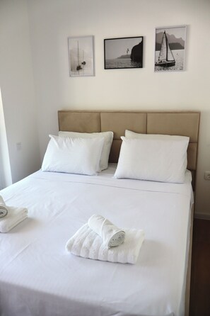 A neatly made bed with white linens and pillows, a folded towel, and three framed pictures on the wall above. - Fighen Apartment (Budva)