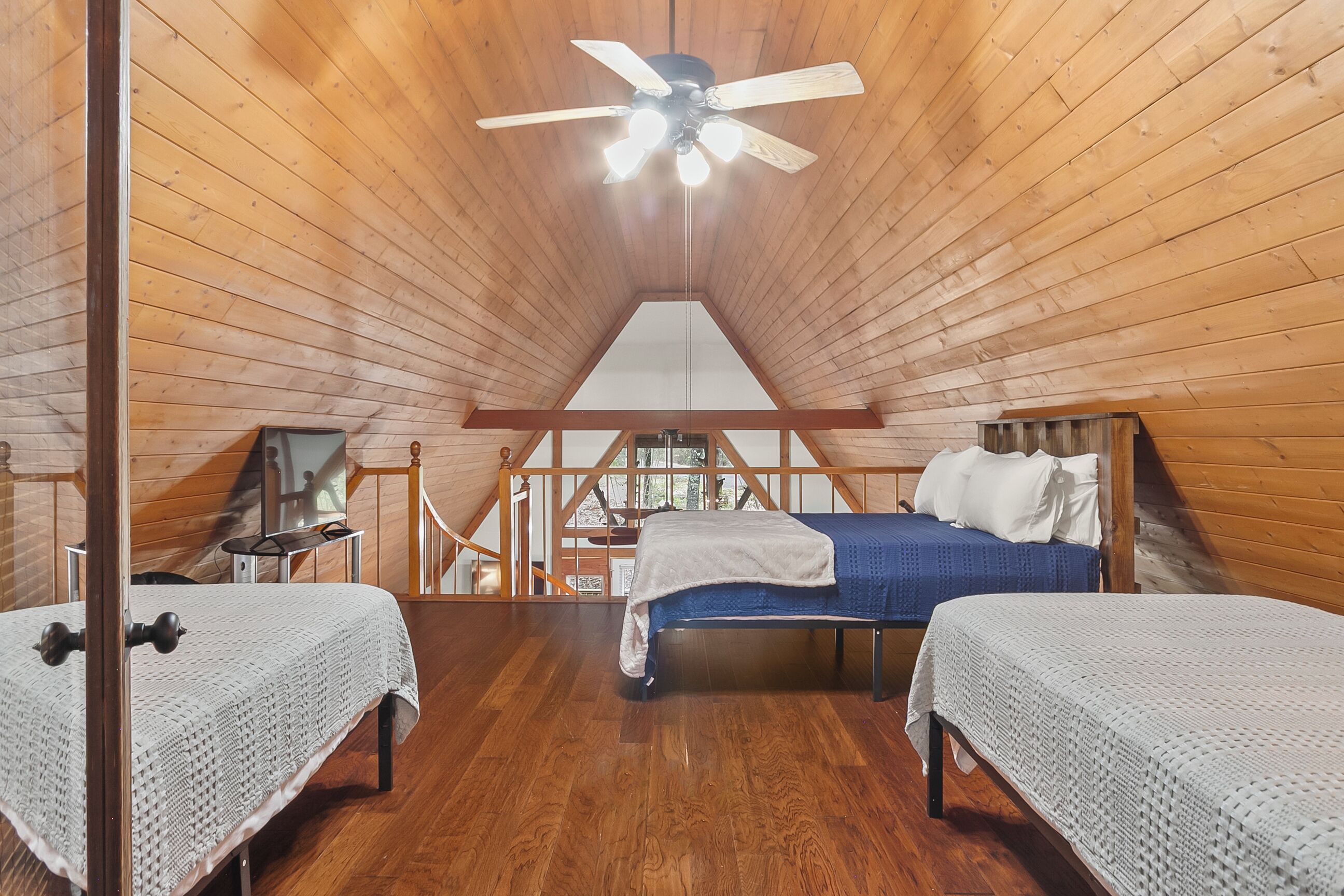 Un dormitorio acogedor en el ático, con paredes de madera, techo inclinado y ventilador de techo. Hay dos camas con ropa de cama blanca y una pequeña mesa con una silla.
