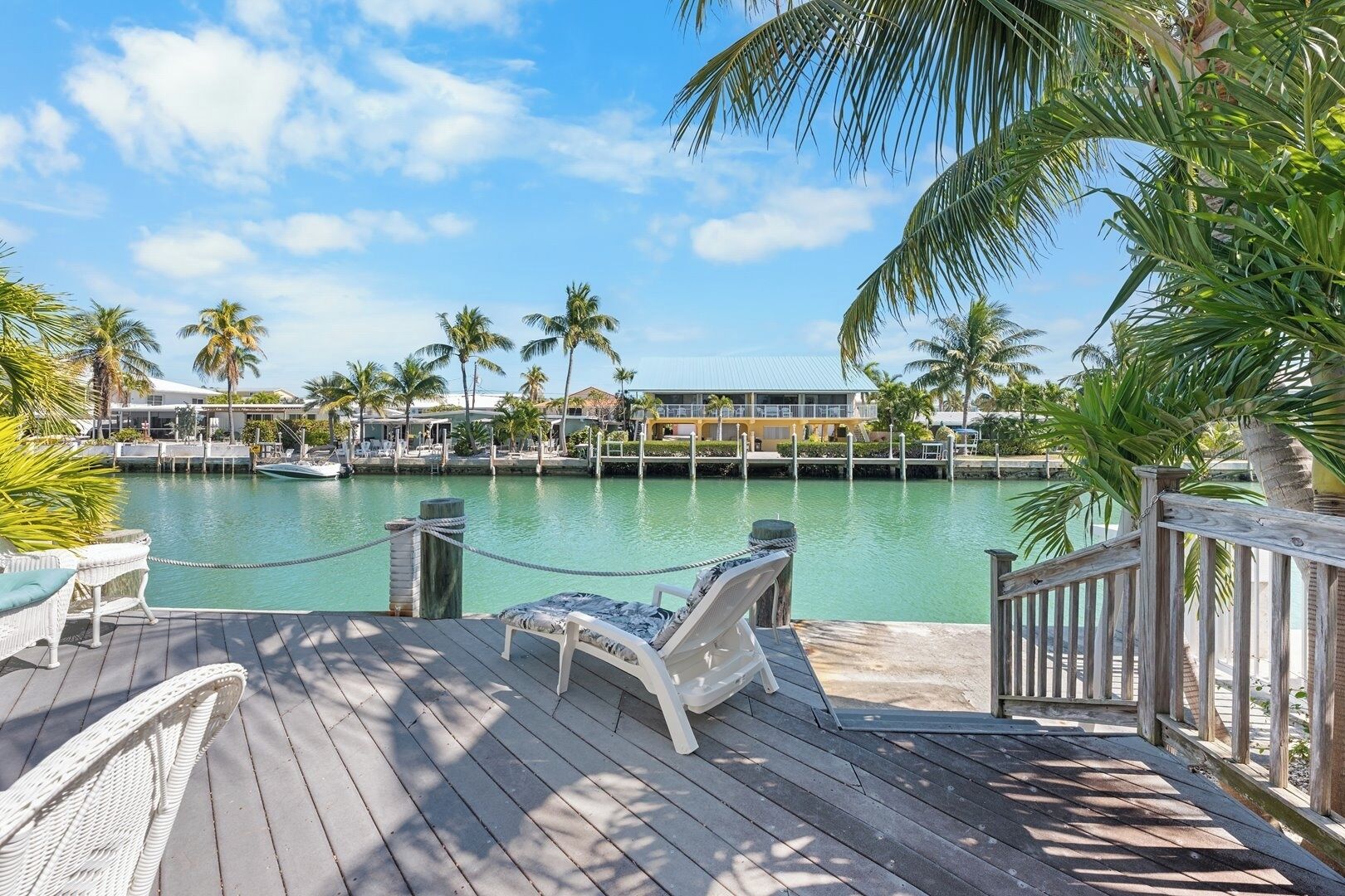 Een houten terras met lounge stoelen met uitzicht op een jachthaven met boten en palmbomen.