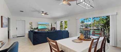 A spacious living area with a large sofa, a dining table with chairs, a ceiling fan, and a view of a pool through large windows.