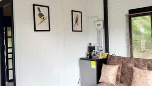 A room with white brick walls, two framed bird illustrations, a grey refrigerator, a brown sofa, and a window with blinds.