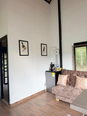 A room with white brick walls, two framed bird illustrations, a grey refrigerator, a brown sofa, and a window with blinds.