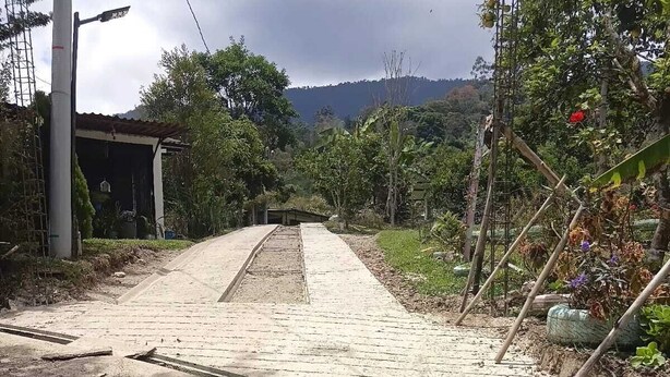 A paved pathway leading to a building with a metal roof, surrounded by vegetation and a hilly landscape.