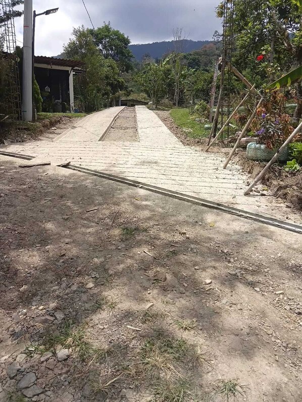 A paved pathway leading to a building with a metal roof, surrounded by vegetation and a hilly landscape.