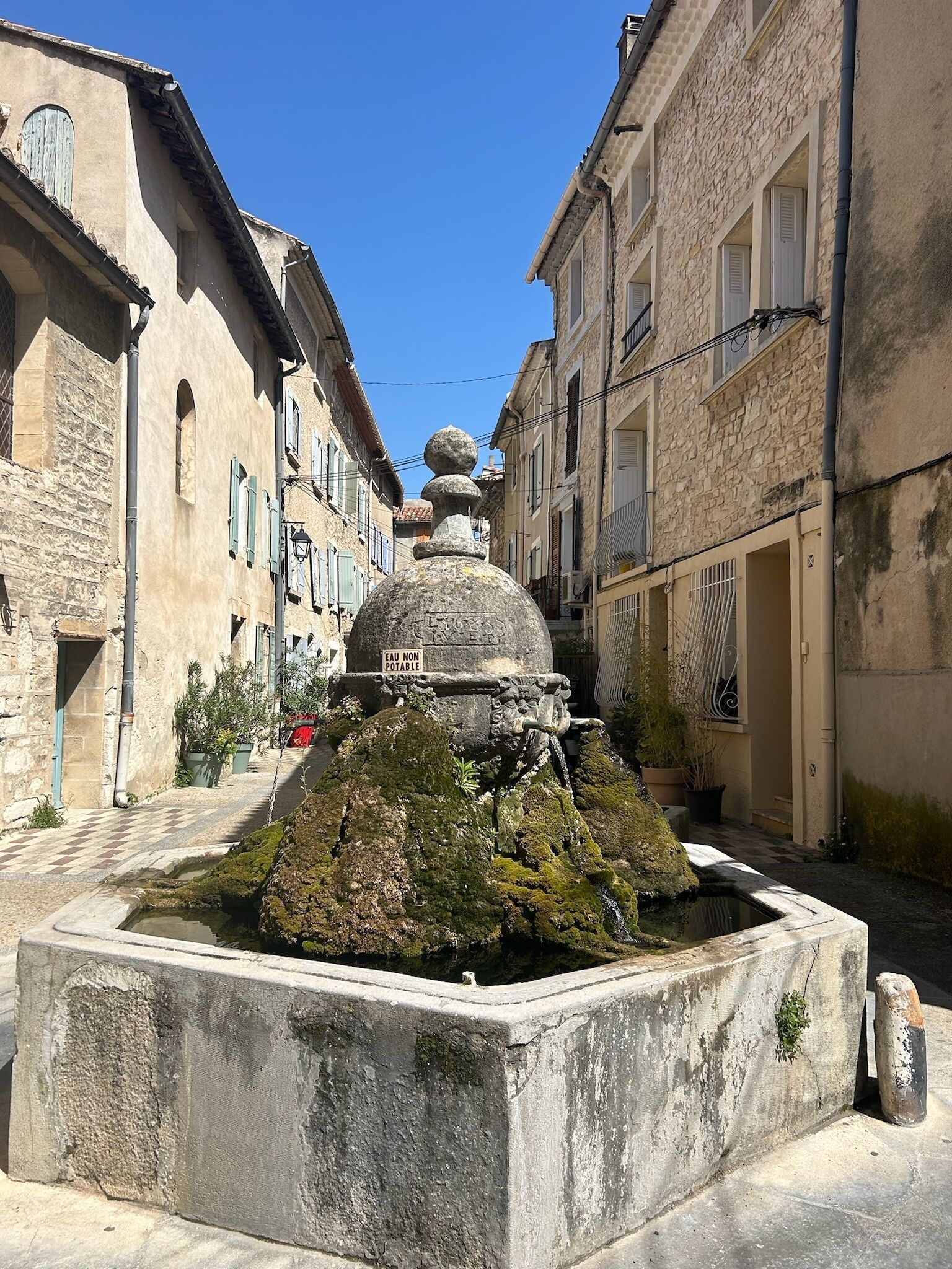 Une fontaine en pierre, recouverte de mousse, se trouve dans une cour entourée de vieux bâtiments.