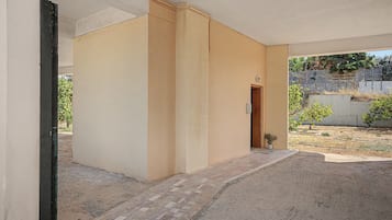 Een overdekt buitengebied met een geplaveid pad dat naar een deur leidt, omgeven door een lage muur en een kleine tuin.