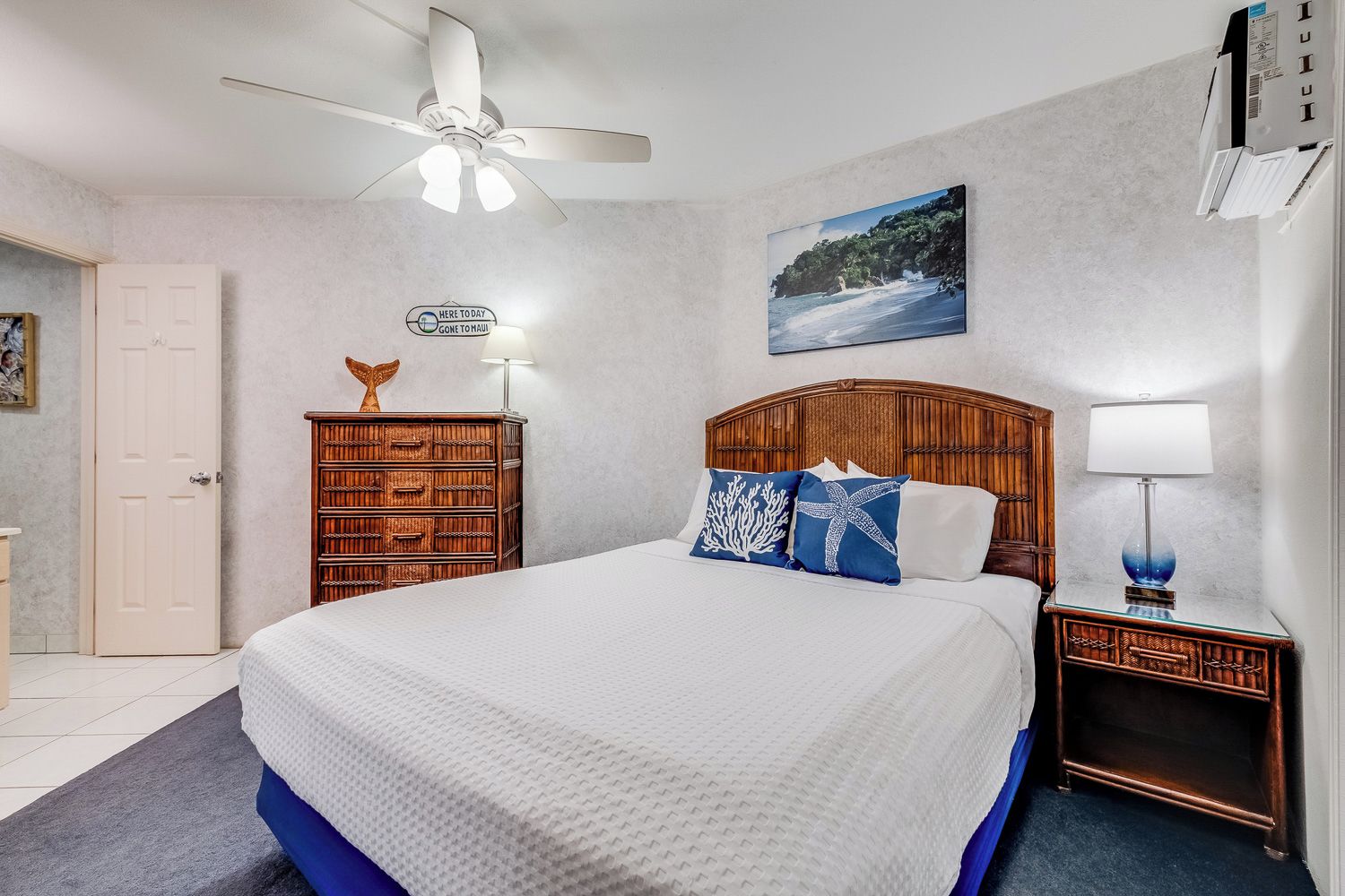 A bedroom with a bed, a wooden headboard, a nightstand with a lamp, a ceiling fan, a door, and a wall-mounted picture.