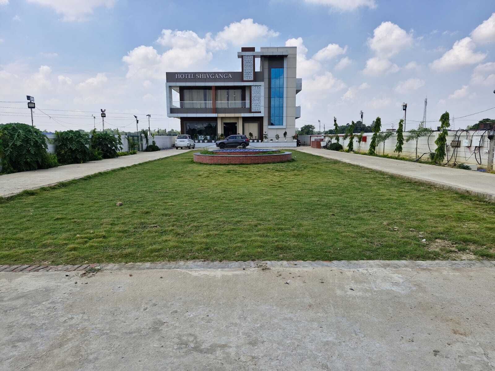 Un moderno edificio de hotel con un letrero que dice 'HOTEL SHIVGANGA' y un césped ajardinado con una fuente circular.