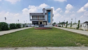 Un moderno edificio de hotel con un letrero que dice 'HOTEL SHIVGANGA' y un césped ajardinado con una fuente circular.