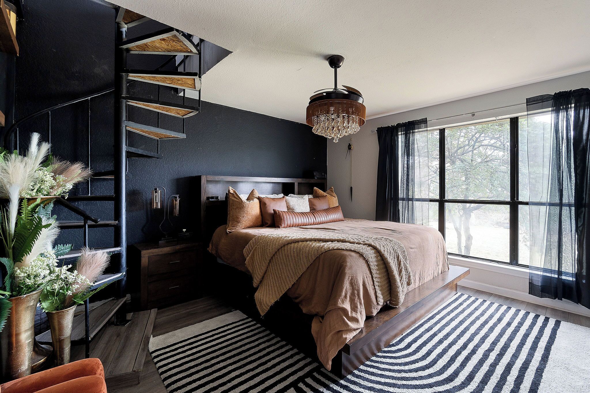 A bedroom with a large bed, a staircase, a chandelier, and a view through a window.