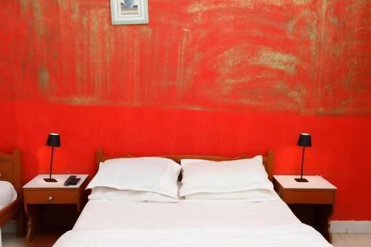 Een bed met witte lakens, twee nachtkastjes met lampen en een rode muur met een ingelijst beeld.