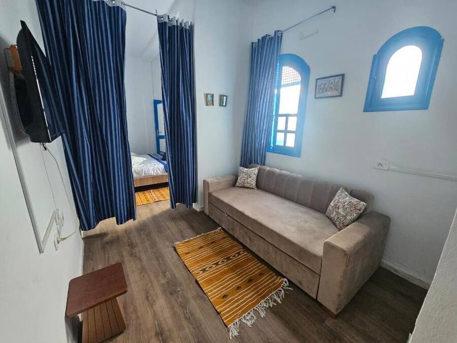 Een kamer met een bank, een houten vloer, een aan de muur bevestigde televisie en een raam met gordijnen.
