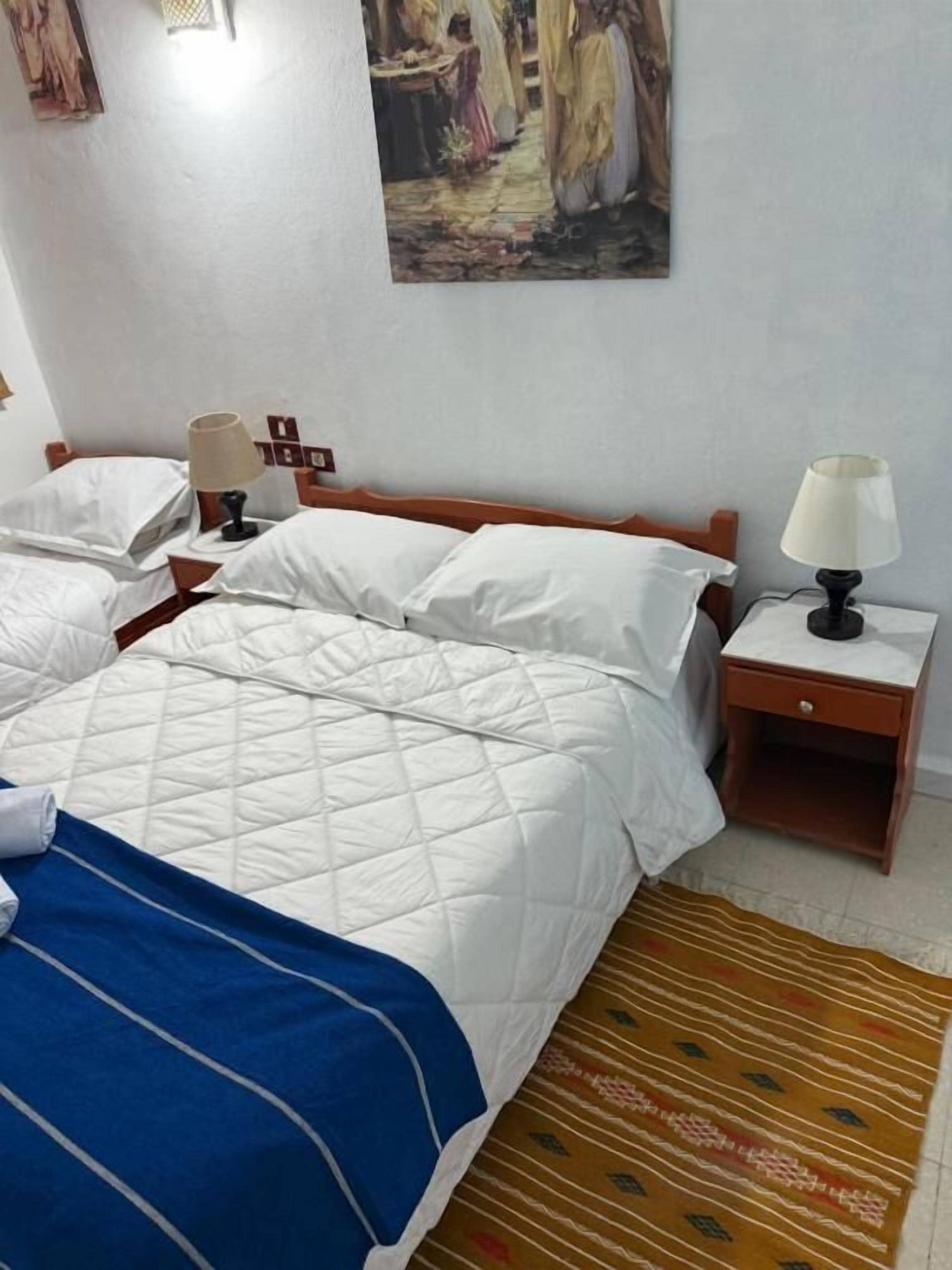 Habitación de hotel con dos camas, una mesita de noche de madera con lámparas, una alfombra estampada y un cuadro en la pared.