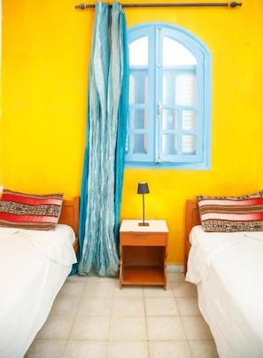 Een kamer met twee bedden, een raam met blauwe luiken, een gordijn, een nachtkastje met een lamp en een betegelde vloer.