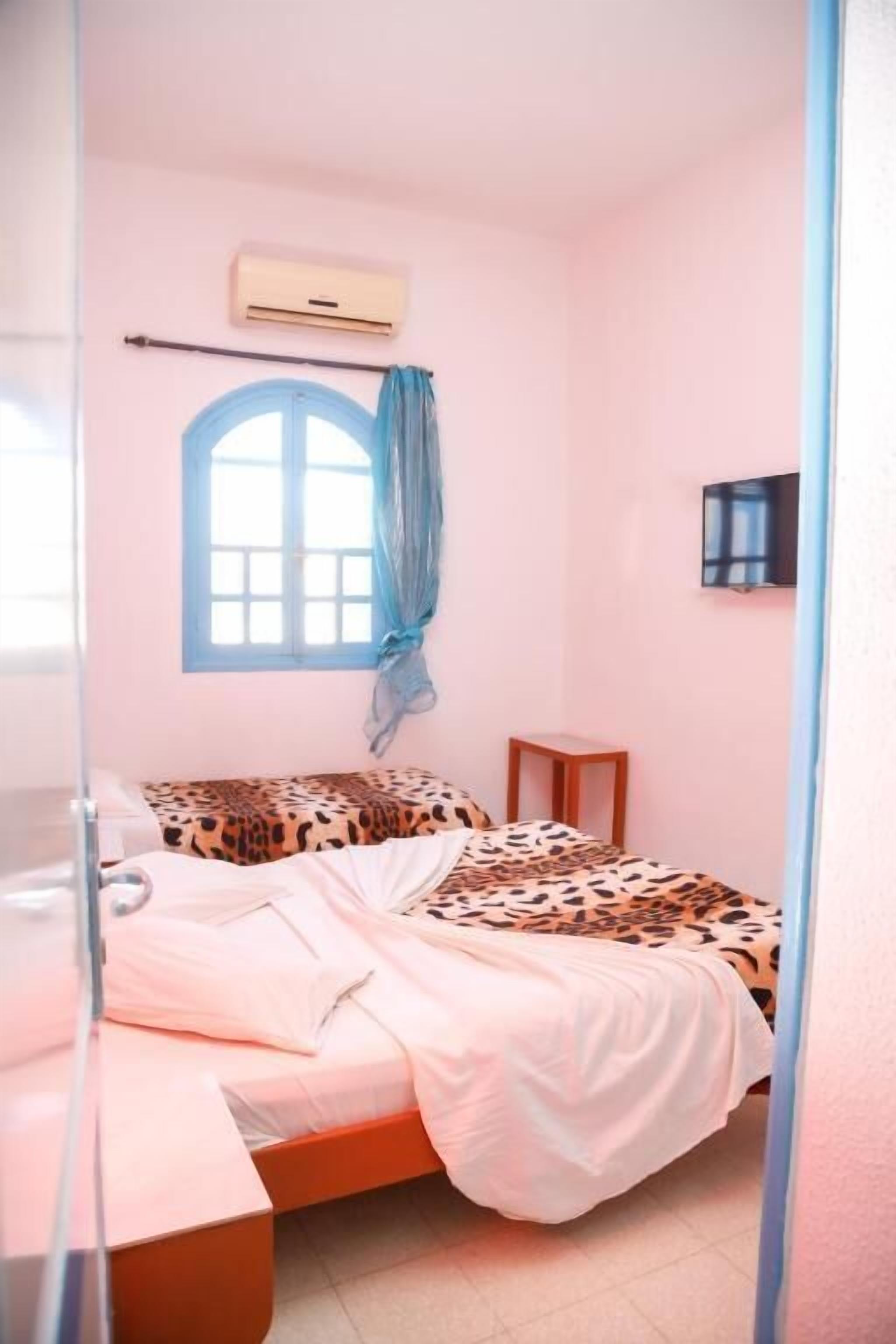 Un dormitorio con dos camas, una con cubrecama estampado de leopardo, una ventana con cortinas azules, un equipo de aire acondicionado y una mesita de noche pequeña.