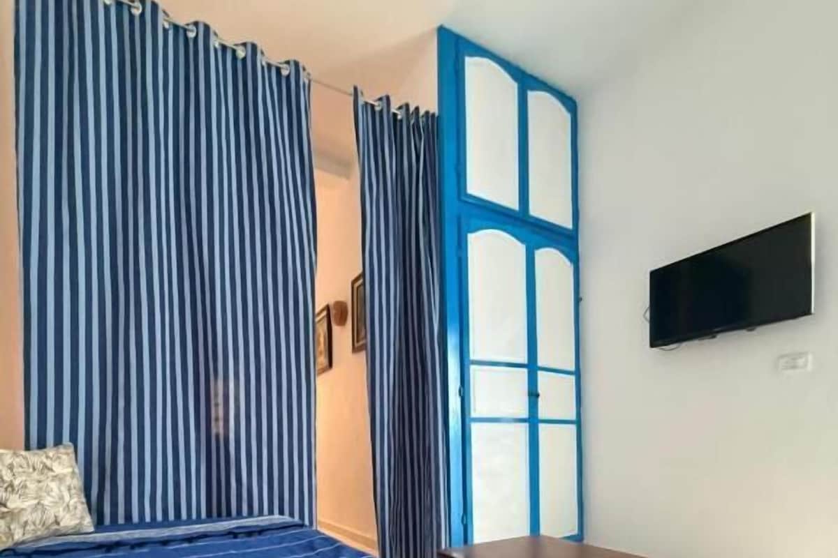 Een slaapkamer met een bed, gordijnen, een televisie en een klein tafeltje.
