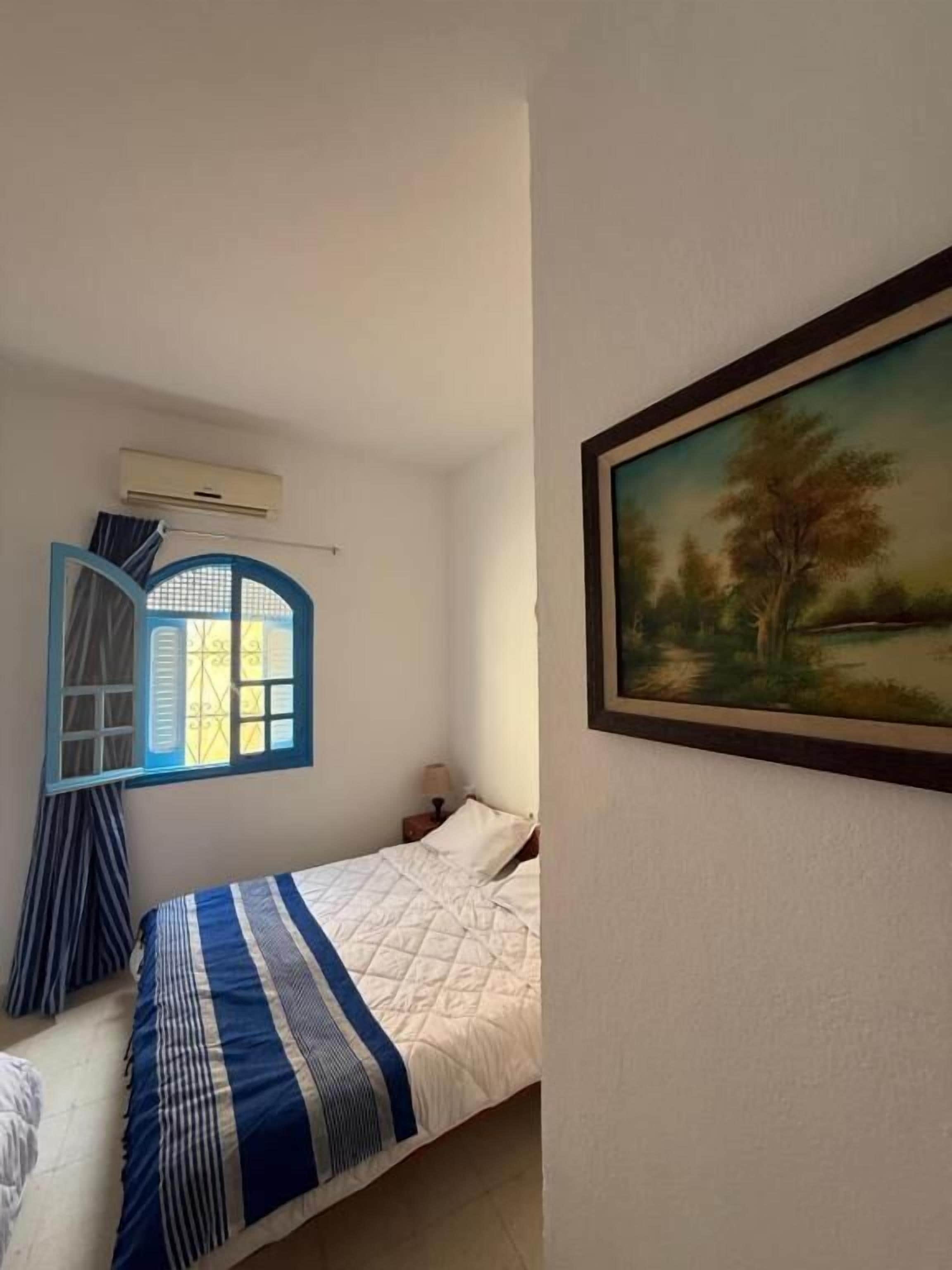 Un dormitorio con una cama, una ventana con cortinas, un equipo de aire acondicionado y un cuadro de un paisaje en la pared.